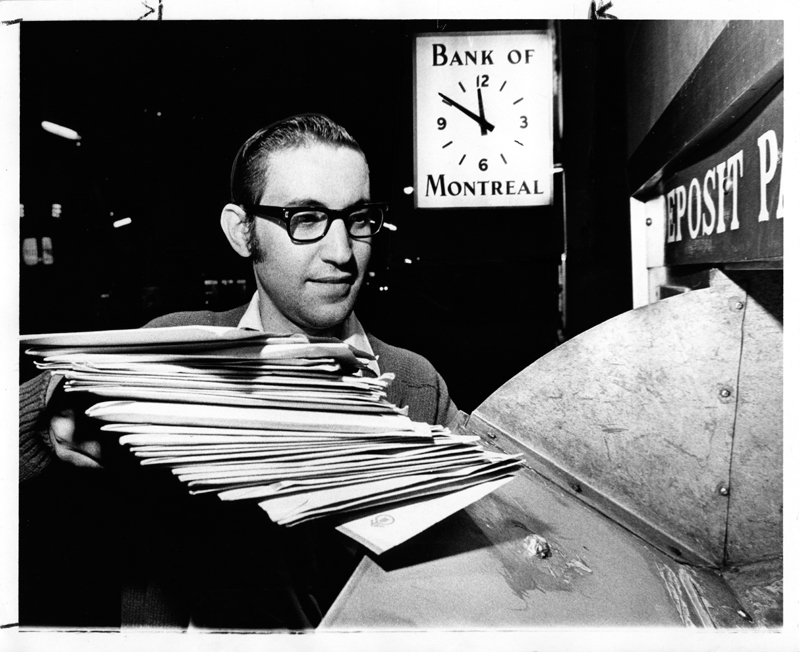 A man in dress shit, pullover sweater and heavy, dark rimmed glasses places a stack of envelopes in a mail slot out side a bank. A large clock featuring the words"Bank of Montreal" is visible in the background.