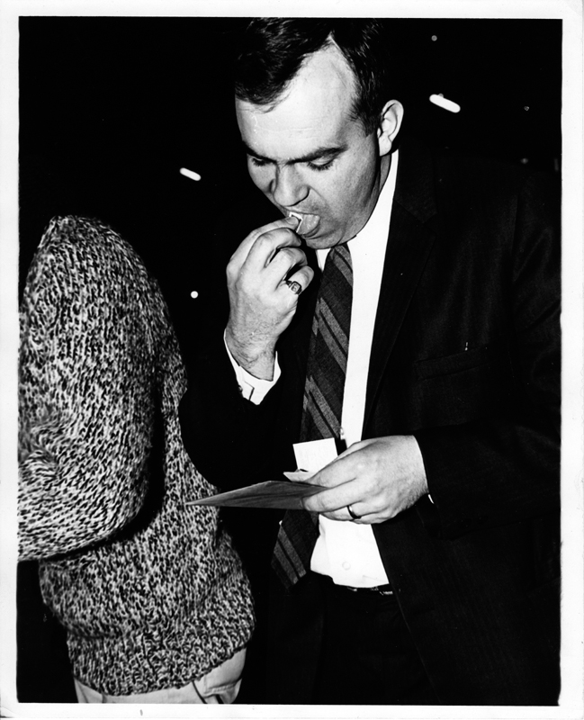 A man in a suit and tie licks a stamp for an envelope he is about to mail. A person is a heavy cable sweater is visible in the background.