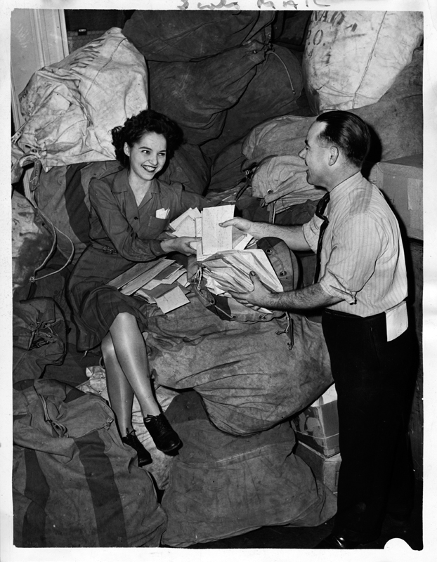 A young woman in a dress is seated on top of a heap of mail bags, posing with a man with a handfull of tax forms.
