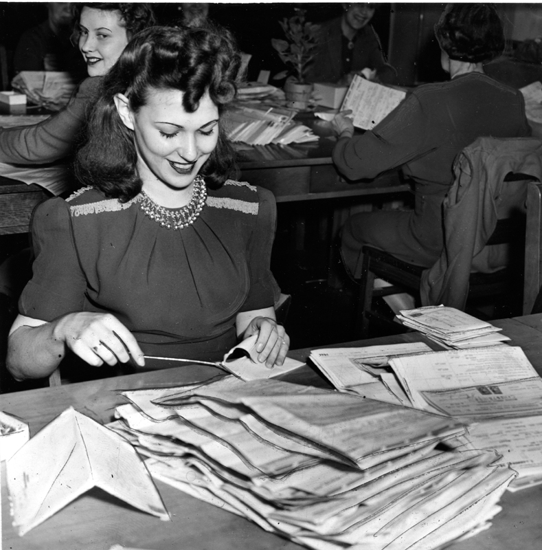A young woman in a dress opens mailed tax forms and piles them before her on a desk. Three women performing similar tasks are visible in the background.