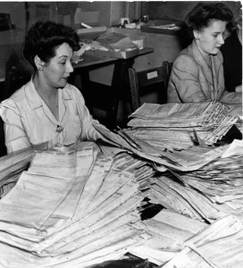 A woman in a light coloured blouse works at a deask piled high with income tax forms. A woman in a suit jacket is visible in the background.