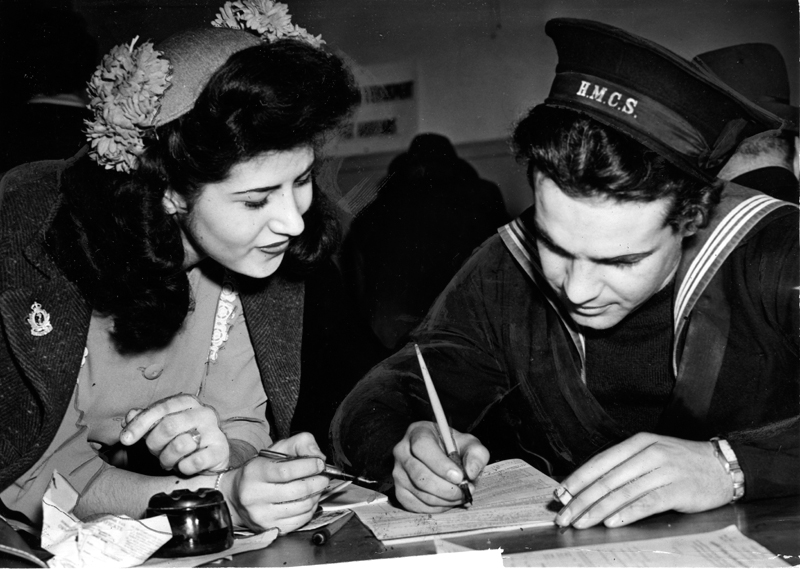 An unidentified H.M.C.S. sailor in uniform filing a tax return while a woman in a flowered hat looks on.
