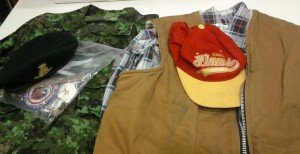 A green military uniform is visible, as is a black military beret and a plastic bag full of badges, pins and medals. Also visible is a flannel checkered shirt, a beige heavy fabric vest and a Calgary Flames baseball cap.