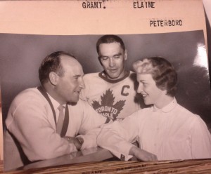 Toronto Maple Leafs coat Billy Reay leans on edge of rink barrier speaking to Elaine Grant, who rests her elbow on the barrier. Captain George Armstrong in his captain's jersey is standing between them..