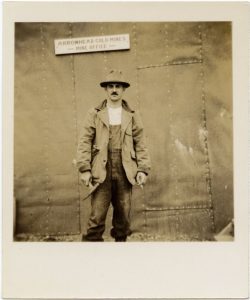 Picture of Arnold Hoffman from ankles up, wearing a hate and jacket, stnading infront of a metal building and sign above him.