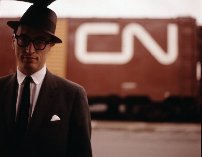 Allan Robb Fleming infront of a Canadian National Railway cart: A man with glasses in a suit and hat, with an umbrella, standing in front of train car with the letters ‘C’ and ‘N’ on it. 
