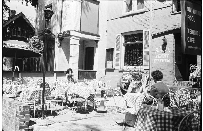 Image of cafe patio. Two individuals sitting at separate tables. Image is of cafe the Penny Farthing which was a popular scene for Yorkville and Toronto's folk music scene.