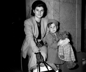 A woman, wearing patterned gloves and holding handles of purse and bag with right hand, sitting on luggage next to child, wearing gloves and pillbox-style hat and holding teddy bear.