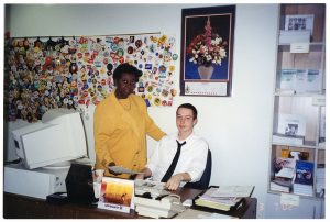 Image of Jean Augustine in yellow suit posing beside young man in white shirt and black tie seated at an office desk. Behind them is a series of wall hangings holding many political and activist buttons.