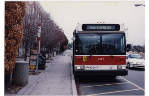 Image of a 106E Express bus standing in front of its stop at the bus loop near York Lanes. Cars driving and a person walking are visible in the background.